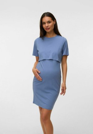 Femme enceinte en robe de maternité bleu clair tenant doucement son ventre, debout devant un fond blanc uni.
