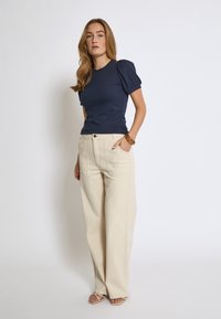 Marinblå ribbad kortärmad topp med puffärmar, ihop med högmidjade beige vida byxor och neutrala sandaler, stående mot en enhetlig bakgrund.