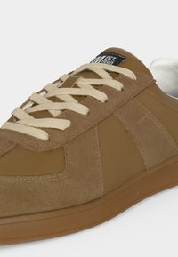 Καφέ σουέντ και δερμάτινο αθλητικό παπούτσι με μπεζ κορδόνια και ετικέτα στη γλώσσα με την ένδειξη "EST 1990", ορατό από πλευρική και εμπρόσθια γωνία.