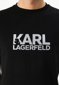 Μαύρο βαμβακερό T-shirt που διαθέτει μια μεγάλη ασημένια γραφική παράσταση με το "KARL LAGERFELD" και μια σιλουέτα προφίλ logo πάνω από το κείμενο.