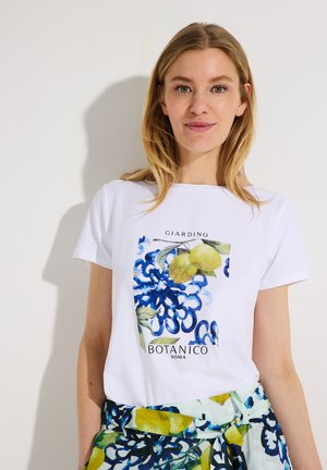 Blonde Frau trägt weißes T-Shirt mit blauem Blumen- und Zitronenmuster, kombiniert mit passendem Rock in Blau, Grün und Gelb mit Muster.