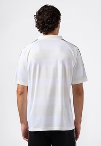 Man met krullend donker haar, gekleed in een witte blouse met subtiele horizontale strepen, met zijn rug naar de camera gekeerd tegen een effen witte achtergrond.
