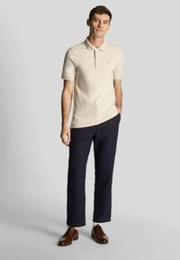 Beiges Poloshirt mit Kragen und kurzen Ärmeln, kombiniert mit marineblauen Hosen und braunen Lederschuhen, versehen mit einem dezenten eingestickten Emblem.