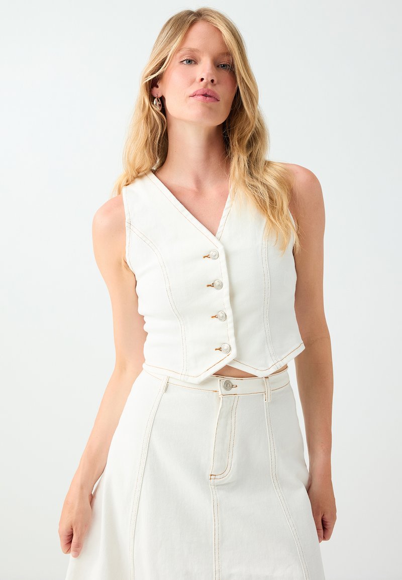 Femme aux longs cheveux blonds portant un gilet blanc sans manches boutonné et une jupe blanche taille haute avec des coutures orange visibles sur un fond uni.