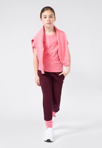 Κορίτσι περπατάει μπροστά φορώντας ροζ φούτερ ριγμένο στους ώμους, κοραλί μπλούζα, μπορντώ φόρμες, ροζ κάλτσες και λευκά αθλητικά παπούτσια.