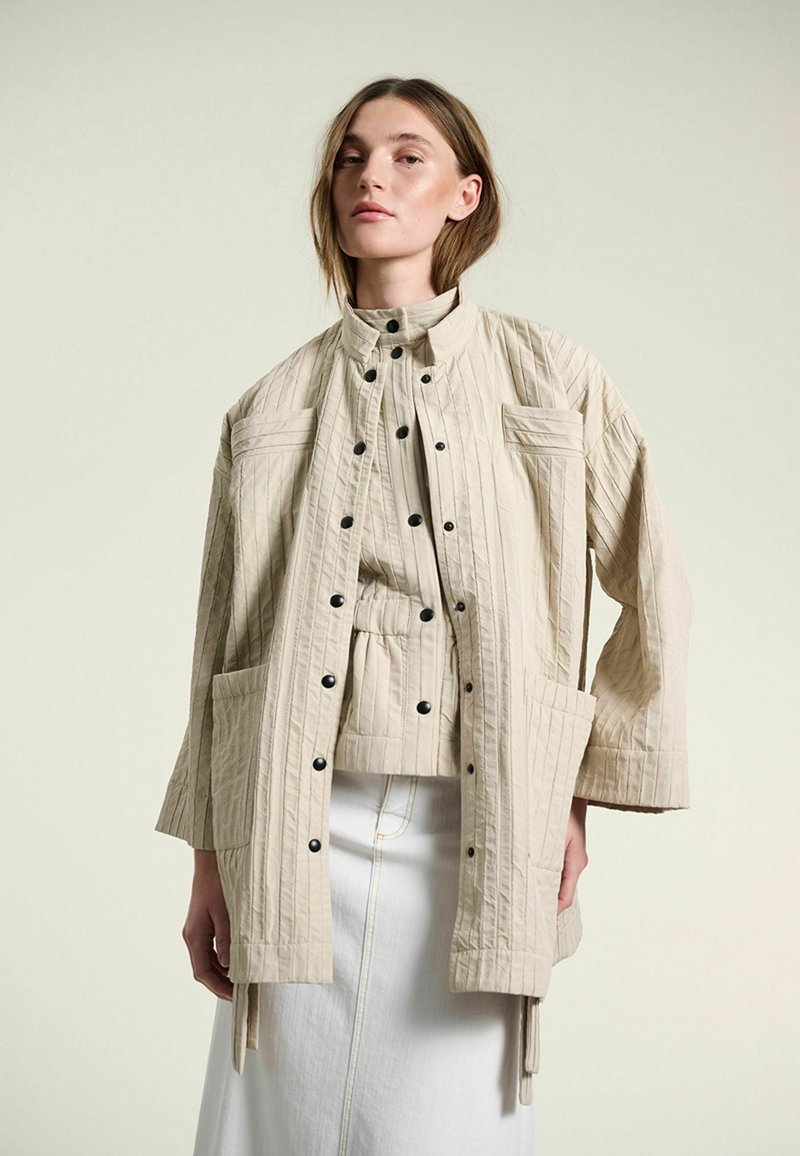 Femme portant une veste matelassée beige boutonnée avec grandes poches par-dessus un haut assorti et une jupe blanche, debout devant un fond uni.