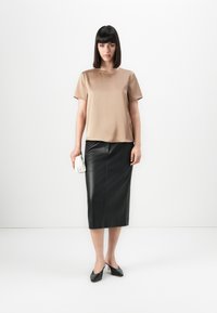 Vrouw die tegen een witte achtergrond staat, gekleed in een beige blouse met korte mouwen, een zwarte leren midi rok, zwarte hakken en een witte clutch vasthoudt.