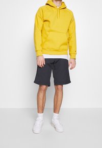 Gul huvtröja med ficka fram, matchad med mörkgrå shorts och vita sneakers. Enkelt mönster, mjuk textur och avslappnad passform.