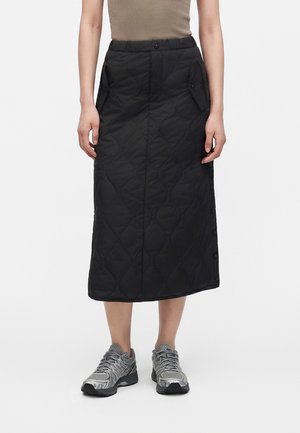 Persona con una falda midi negra acolchada con botones laterales, top sin mangas color topo y zapatillas deportivas grises, de pie sobre un fondo blanco.
