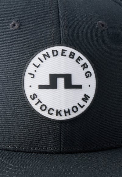 Μαύρο καπέλο με έναν κυκλικό λευκό πάτο μπροστά που απεικονίζει ένα μαύρο γεωμετρικό λογότυπο και το κείμενο "J. LINDEBERG STOCKHOLM" κατά μήκος του άκρου.
