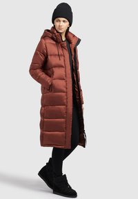 Long, padded coat in dark rust color with a hood, zip front, and two side pockets. Styled with black leggings and textured black boots.