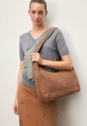 Młoda kobieta trzymająca dużą brązową torbę na ramię o teksturowanej powierzchni, ubrana w szarą bluzkę z krótkim rękawem, brązową spódnicę z zdobieniami oraz zielony sweter zarzucony na ramiona.