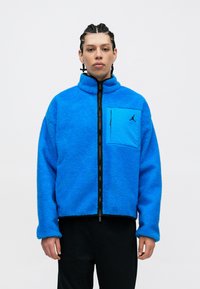 Niebieska polarowa kurtka o strukturalnym designie, z wysokim kołnierzem, pełnym zamkiem błyskawicznym oraz kieszenią z przodu z detalem logo.