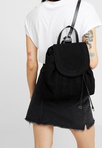 Osoba v bílém tričku a černé denimové sukni, která na zádech nese černý semišový batoh se šňůrkovým uzávěrem.