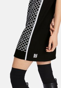 Schwarzes Kleid mit einem gemusterten weißen Seitenteil, kontrastierenden weißen Akzenten und einem Logodetail am Saum. Das Material wirkt glatt und gestrickt.