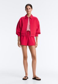 Femme portant une veste rouge zippée, un short rouge assorti avec cordon, une chemise rouge et blanche rayée, et des sandales à plateforme noires, debout.
