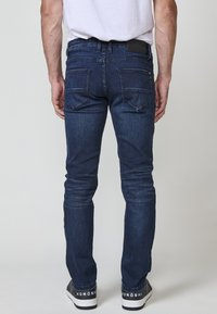 Muškarac u tamnoplavim slim fit farmerkama i crnim tenisicama s bijelim potplatima, stoji okrenut prema jednostavnoj svijetlosivoj pozadini.