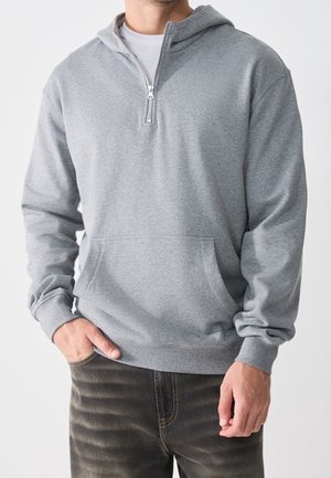 Homme portant un sweat à capuche gris clair à demi-fermeture éclair avec poche avant et jean foncé délavé, main dans la poche, sur fond uni.