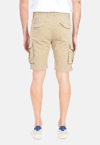 Pantaloni cargo beige con molte tasche laterali e tasche posteriori, realizzati in cotone. Indossati con sneaker bianche con un accento blu.