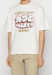 Muž oblečený v bílém oversized tričku s barevným japonským textem a grafikou, v kombinaci s béžovými kalhotami.
