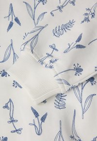 Valkoinen kangas sinisellä kasviaihellemalla, jossa on erilaisia kasveja. Kuvassa näkyy purjeribadilla varustettu yksityiskohta. Pehmeä rakenne.