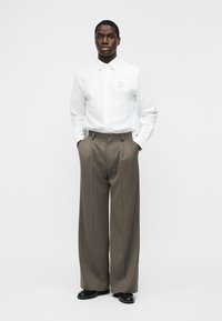 Moški stoji z rokami v žepih, oblečen v belo srajco z gumbi in logotipom "EA" ter v rjavih širokih hlačah in črnih čevljih.