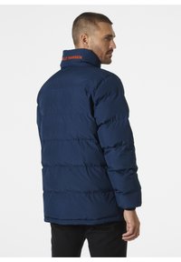 Chaqueta acolchada azul marino con cuello alto, que presenta paneles horizontales cosidos y un logo naranja de "Helly Hansen" en el cuello.