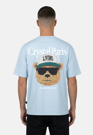 Uomo che indossa una maglietta azzurro chiaro con grafica di orso con occhiali da sole e cappellino bianco, scritte "Crystal Paris" e "Living Life" sul retro.