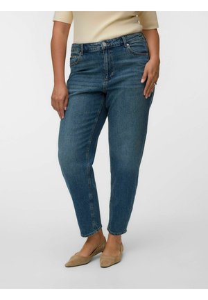 Femme portant un jean bleu slim taille haute, un haut beige à manches courtes et des ballerines pointues marron clair sur un fond uni.
