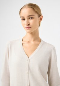 Svetle béžový svetr s V-výstřihem, texturovaným pleteným designem a pěti knoflíky vpředu. Má dlouhé rukávy a měkký, hladký povrch.