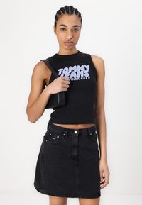 Svart avkortet tanktop med "TOMMY JEANS NEW YORK CITY" trykk, parret med et høyhaltet svart denimskjørt. Modellen bærer en liten svart veske.