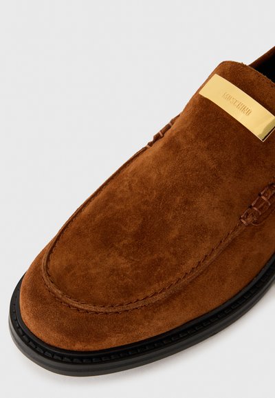 Sapatos mocassim de camurça castanha com biqueira arredondada, sola de borracha preta e um detalhe metálico dourado na parte superior com gravação do nome da marca.