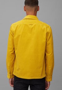 Camisa de pana en amarillo mostaza con cuello abotonado, mangas largas y dobladillo recto. Presenta canaletas verticales y puños con botones.