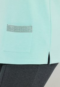 Jasnoturkusowy top z ukończonym brzegiem i bocznymi rozcięciami, z kieszenią ozdobioną małymi srebrnymi kryształkami. Szare legginsy na spodzie.