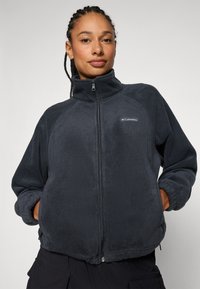 Giacca in pile grigia scura con colletto alto, chiusura con zip e polsini elasticizzati. Presenta una discreta patch del logo sul lato sinistro del petto.