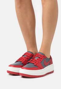 Pieds portant des baskets Nike rouges et grises avec de grosses semelles blanches, se tenant sur un fond blanc.
