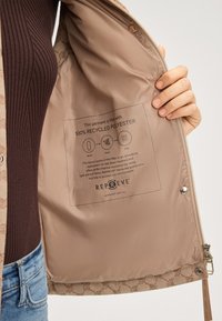 Beige Jackeninnenfutter mit einem Etikett, auf dem "100 % Recyceltes Polyester" steht. Weiche Textur, Reißverschluss und cremefarbene Akzente am Futter.