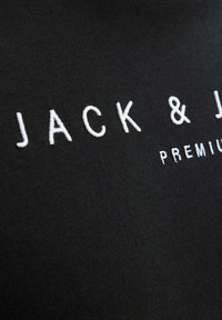 Artimas juodo audinio vaizdas su balta siuvinėta tekstu "JACK & J" ir matoma dalis žodžio "PREMIUM".