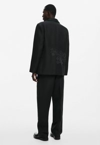 Fekete, személyre szabott öltöny, enyhén túlgondolt blézerrel, amelyen finom virágmintázat található a hátsó részen, bő szárú nadrággal párosítva.