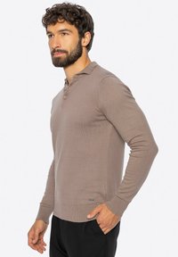 Mężczyzna z brodą w beżowej koszulce polo z długim rękawem i czarnych spodniach, stojący z jedną ręką w kieszeni, patrzący na bok.