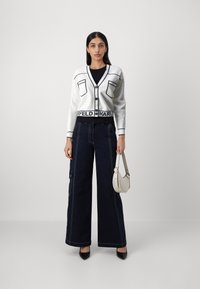 Vitt kort kardigan med svarta fickkantlinjer, kombinerad med mörka vida jeans med ljusa sömmar. Modellen håller en liten vit väska.