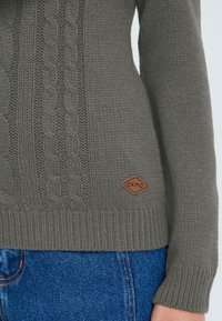 Gray knitted sweater with cable patterns, ribbed cuffs and hem, featuring a small brown branded tag at the bottom. Worn with blue denim.