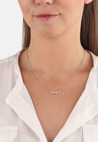 Smycke i silverton med fem runda, klara stenaccenter i en böjd formation, bärs av en person med en vit blus.