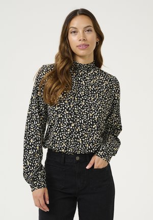 Blouse noire avec col montant, manches longues, présentant un motif abstrait crème et or éparpillé. Associée à un jean noir taille haute.