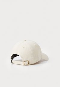 Beige bomullsbasebollkeps med en böjd skärm, justerbart strap, och sex ventilationshål på toppen. Slät textur, avslappnad design.