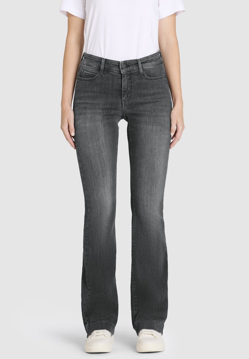 Mujer con jeans bootcut negros desgastados, camiseta blanca y zapatillas blancas, de pie contra un fondo liso de color gris claro.