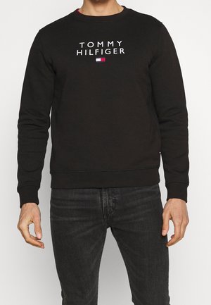 Person trägt einen schwarzen Tommy Hilfiger Sweatshirt mit weißem Logotext und kleinem Flaggenemblem, kombiniert mit dunklen Jeans, vor hellem Hintergrund.