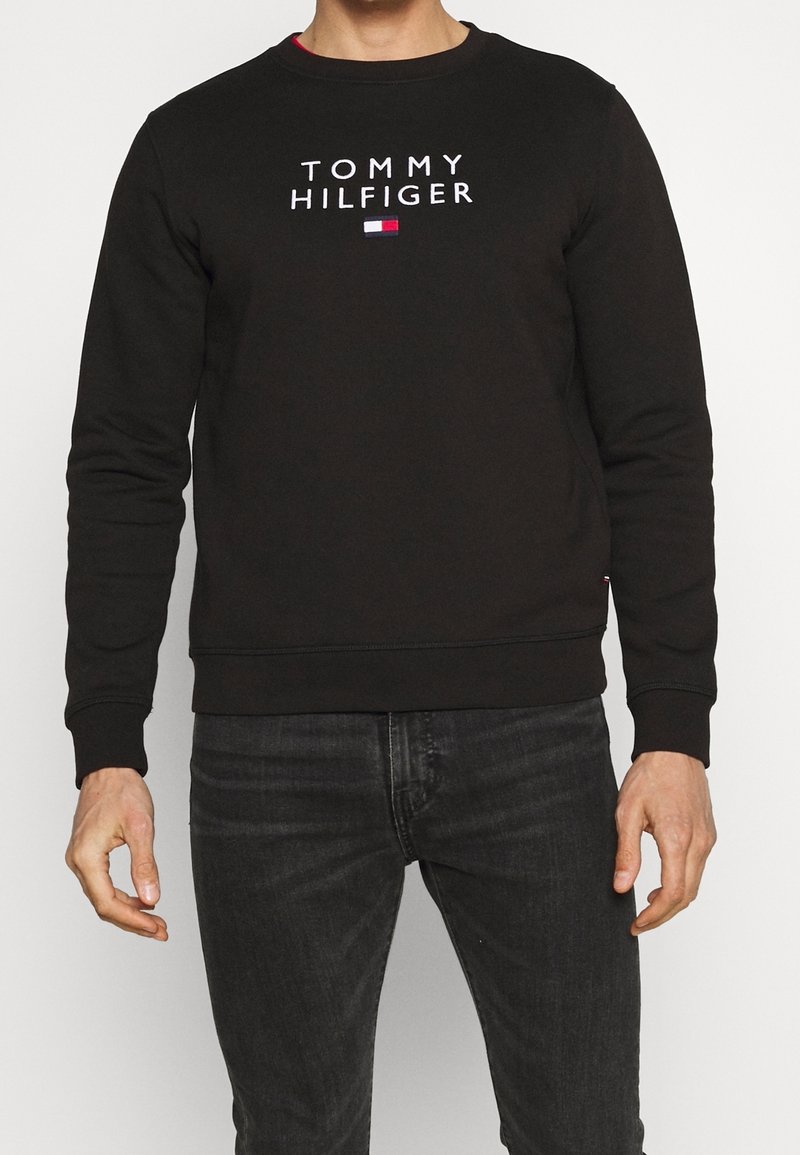 Personne portant un sweat-shirt noir Tommy Hilfiger avec un texte de logo blanc et un petit emblème de drapeau, associé à un jean foncé, sur un fond clair.