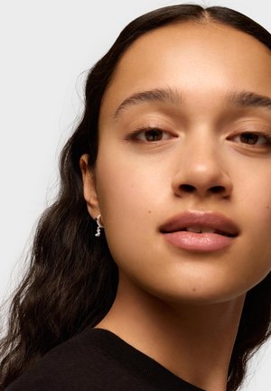 Pendientes de plata con un diseño retorcido y acentos de perla. La modelo tiene el cabello oscuro y ondulado y lleva una blusa negra. El fondo es neutro.