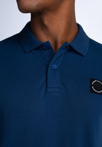 Námořnická modrá polo tričko vyrobené z texturované látky, s dvěma knoflíky na límci a černou nášivkou s kruhovým designem na levém prsu.
